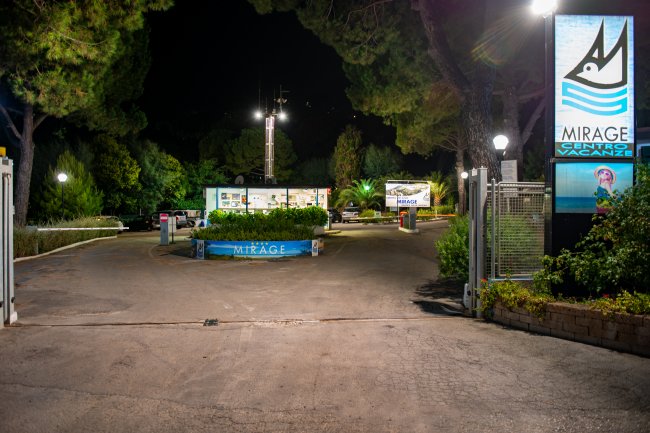 Entrata notturna di un centro vacanze, con luci e cartelloni illuminati tra alberi.