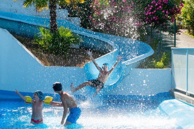 Persone si divertono in piscina con scivolo e spruzzi d'acqua.
