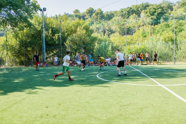 Partita di calcetto su un campo circondato da alberi e spettatori.