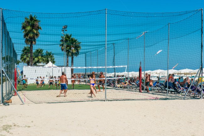 Persone giocano a beach volley su sabbia con sfondo di ombrelloni e palma.
