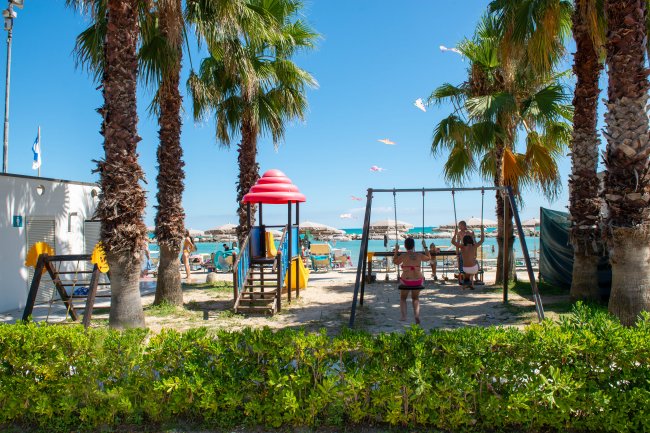 Area giochi con altalene e scivoli tra palme vicino alla spiaggia.