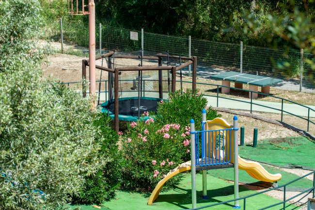 Area giochi all'aperto con scivolo giallo, rete per arrampicata e tavolo da ping pong.
