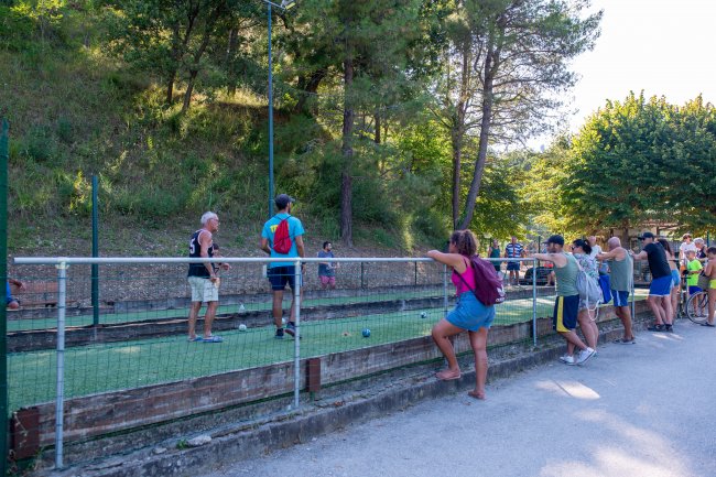 Persone giocano a bocce su un campo circondato da alberi; spettatori appoggiati alla recinzione.