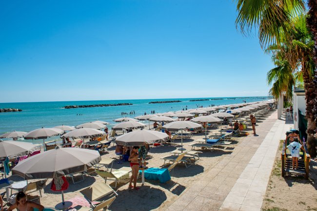 Spiaggia con ombrelloni e lettini lungo la costa, mare azzurro sullo sfondo.