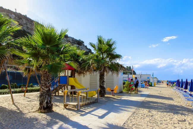 Spiaggia con palme, area giochi per bambini e file di ombrelloni.
