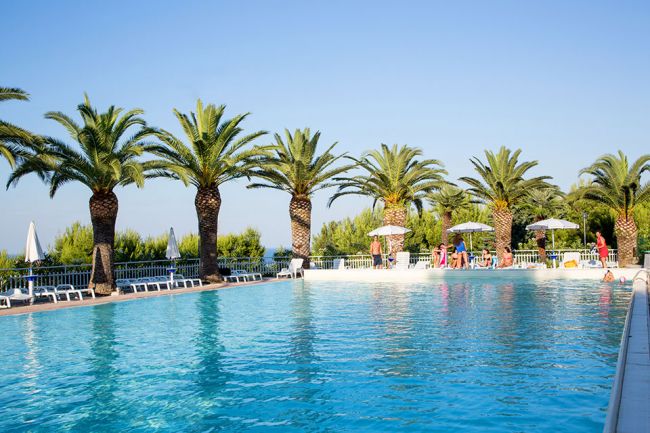 Piscina all'aperto con palme e lettini, cielo blu.