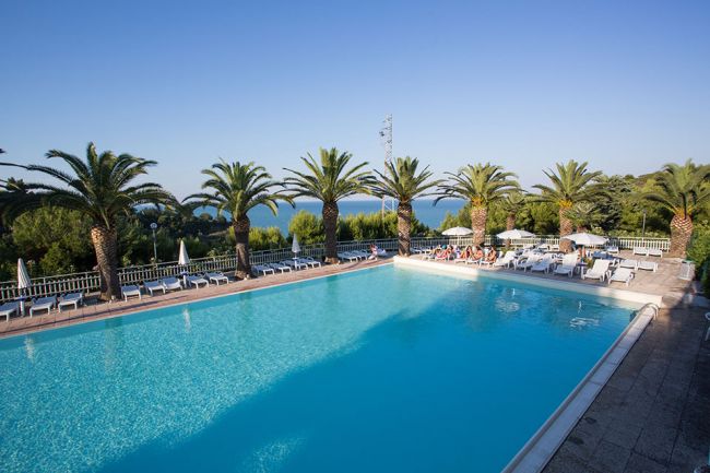 Una grande piscina circondata da palme e sedie a sdraio, con vista sul mare in lontananza.