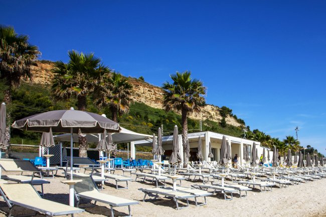 Spiaggia con ombrelloni e lettini sotto un cielo blu, fiancheggiata da palme e una struttura bianca.