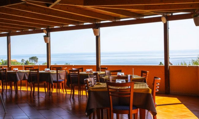 Ristorante con tavoli e sedie in legno, vista mare attraverso un'ampia apertura.