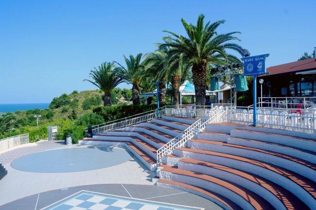 Un anfiteatro all'aperto con palme, sedute vuote e vista sul mare.