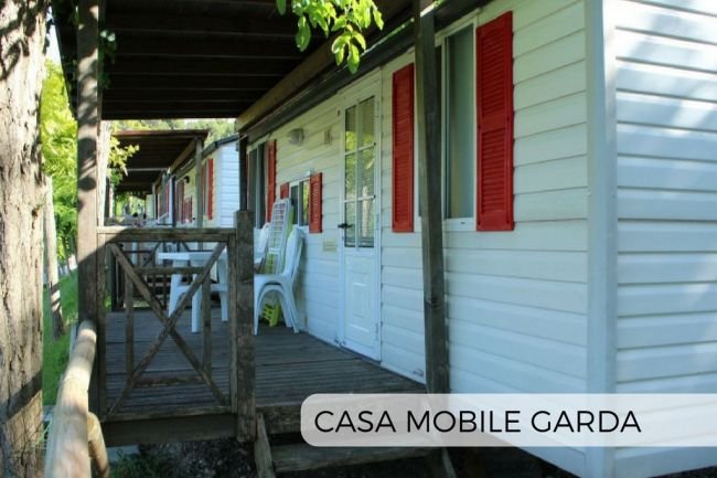 Casa mobile bianca con persiane rosse e veranda in legno, circondata da alberi.