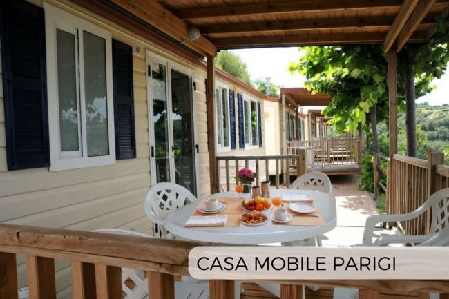 Casa mobile beige con veranda in legno, tavolo apparecchiato per colazione, vista verde.