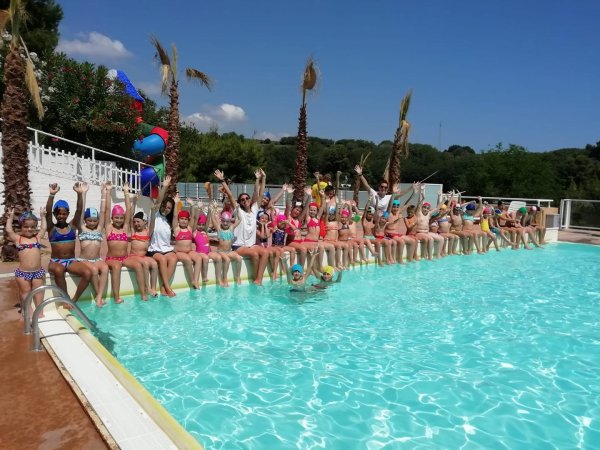 Bambini seduti sul bordo di una piscina alzando le braccia.
