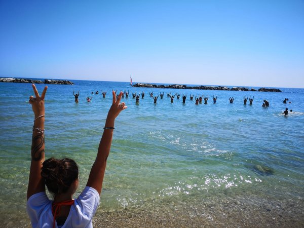 Persona sulla spiaggia con braccia alzate, gente in mare.