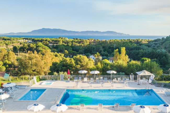 Vista di una piscina con ombrelloni bianchi, circondata da alberi e colline in lontananza.