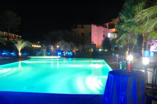 Piscina illuminata di notte con edifici sullo sfondo.