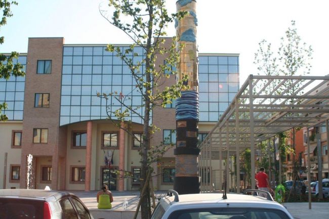 Edificio moderno con facciata in vetro e una scultura alta al centro, circondato da alberi.