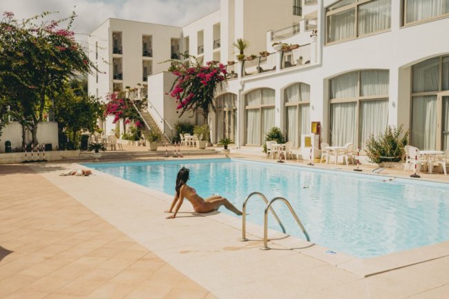 Piscina esterna con persona rilassata a bordo, edificio bianco sullo sfondo.