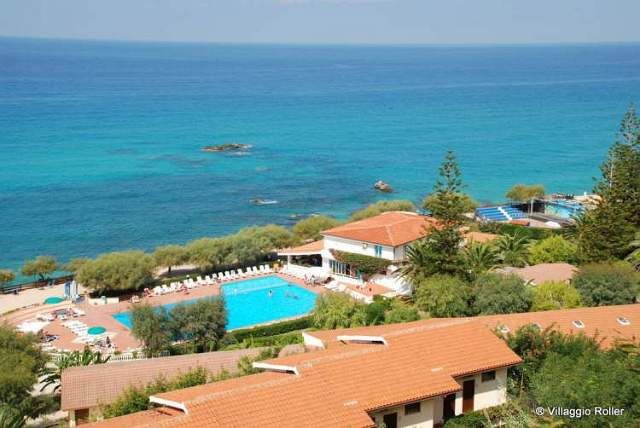Vista su un villaggio turistico con piscina, alberi e mare cristallino.