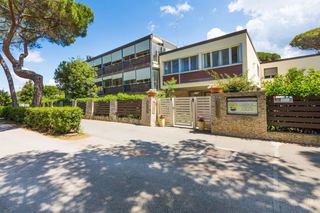 Edificio moderno con cancello e siepi, cielo azzurro e alberi intorno.