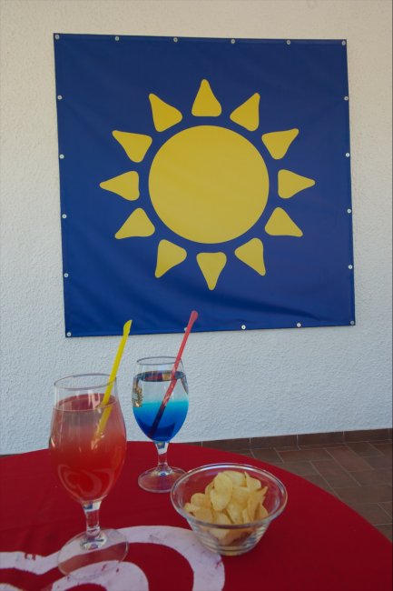 Tavolo con cocktail colorati e patatine. Sfondo con un drappo blu e sole giallo.