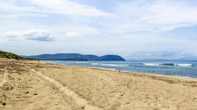 Spiaggia sabbiosa con mare calmo e colline sullo sfondo.