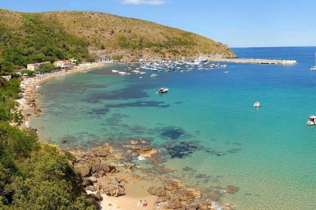 Spiaggia rocciosa con acqua turchese e barche ormeggiate.