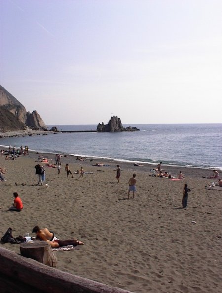Spiaggia affollata con mare e rocce in lontananza.