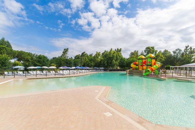 Una piscina con scivolo colorato e aree prendisole, alberi intorno e cielo nuvoloso.
