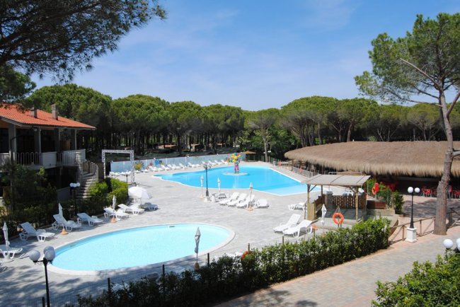 Area piscina con due vasche, ombrelloni bianchi, lettini e un edificio con tetto di paglia.