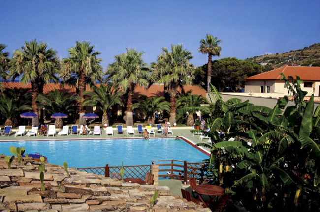 Piscina circondata da palme con sedie a sdraio e ombrelloni blu.