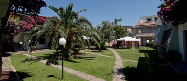 Giardino con palma, vialetti e gazebo; circondato da edifici e fiori.