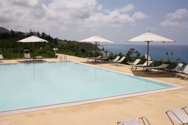 Piscina con ombrelloni, circondata dal verde e vista mare, cielo parzialmente nuvoloso.