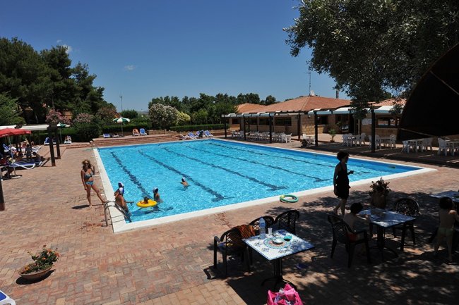 Piscina all'aperto con persone che nuotano e tavolini accanto.