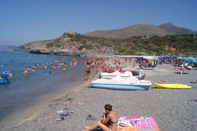 Spiaggia affollata con mare, bagnanti e pedalò sulla riva.
