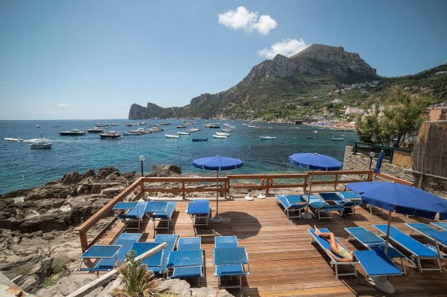 Terrazza con lettini blu, mare e barche in lontananza.