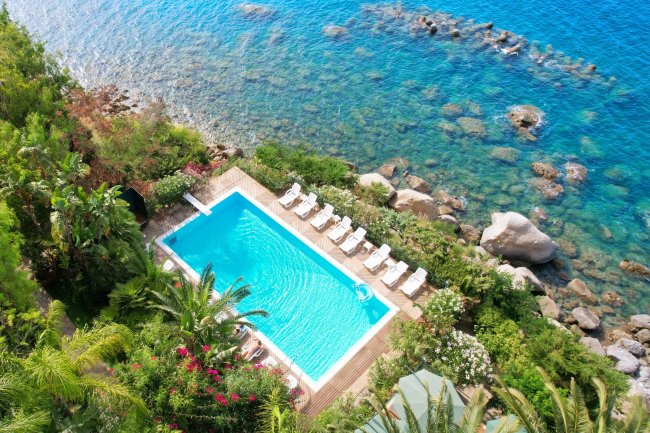 Piscina con lettini, circondata da piante e mare.