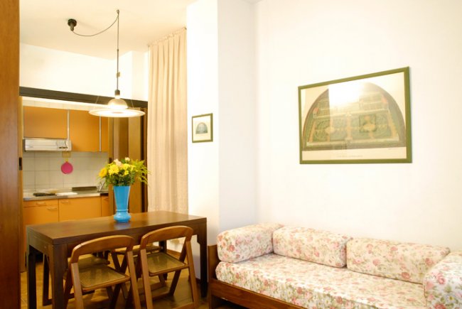 Sala con divano floreale, tavolo in legno, cucina a vista e lampada.