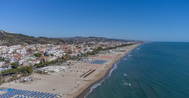 Vista aerea di una spiaggia con ombrelloni e città sullo sfondo.