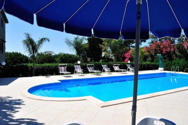 Piscina all'aperto con lettini e ombrellone blu, circondata da palme.