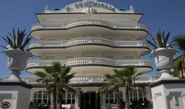 Edificio bianco con balconi curvi e piante davanti.