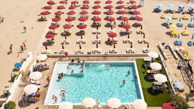 Spiaggia affollata con ombrelloni rossi e piscina.