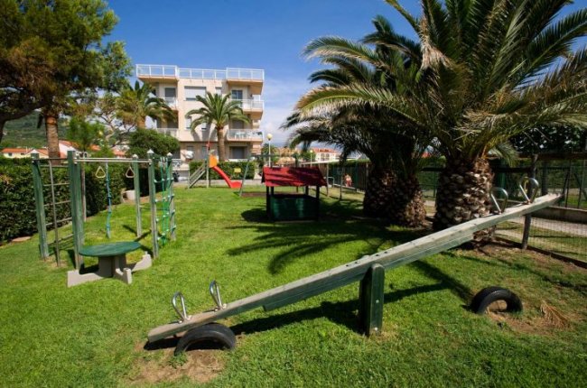 Parco giochi con altalene e scivolo, alberi di palme intorno.