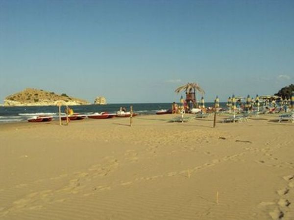 Spiaggia sabbiosa con ombrelloni e pedalò, isola all'orizzonte.