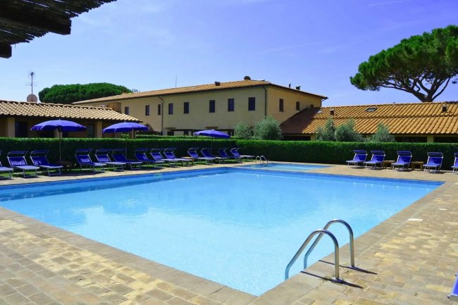Piscina con lettini e ombrelloni blu, edificio sullo sfondo.