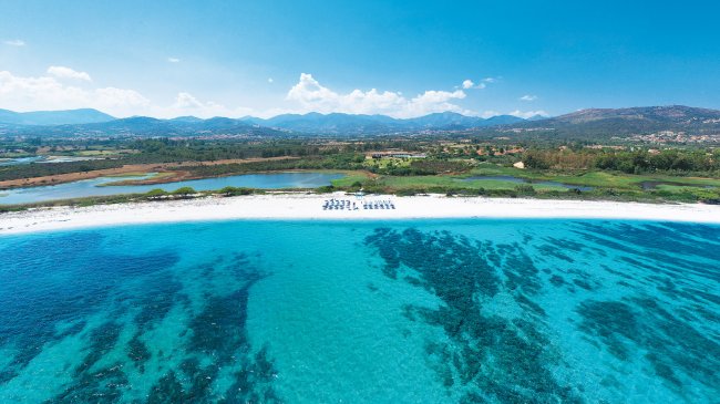 Spiaggia con acqua cristallina e ombrelloni, montagne sullo sfondo.
