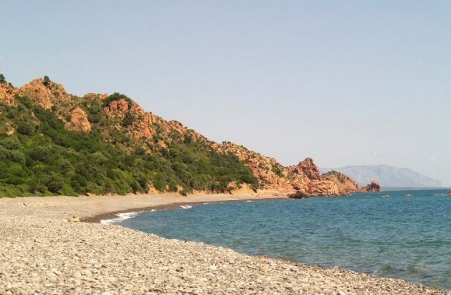 Una spiaggia di ciottoli con mare azzurro e colline verdi in lontananza.