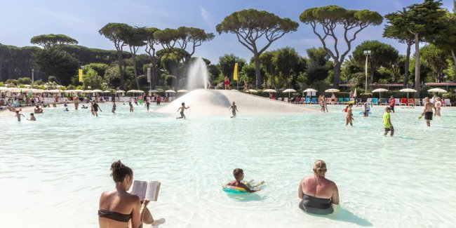 Persone in piscina con baldacchini e alberi sullo sfondo.