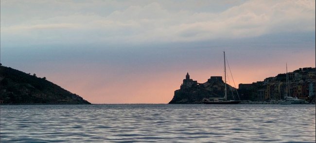 Lungomare al tramonto con barche e colline all'orizzonte.