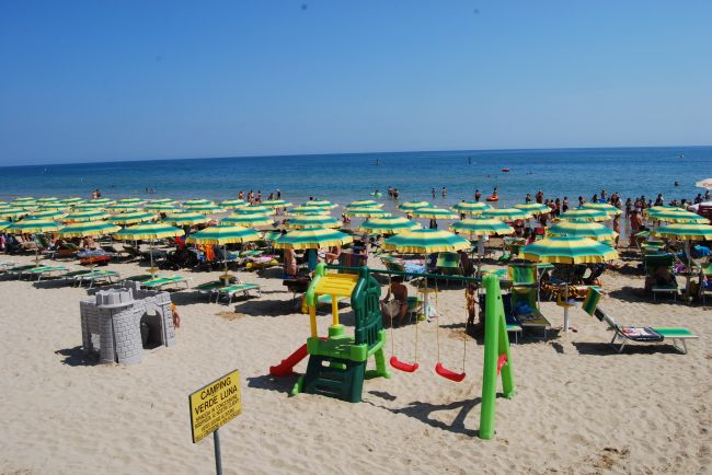 Spiaggia affollata con ombrelloni verdi e gialli, area giochi per bambini e mare sullo sfondo.